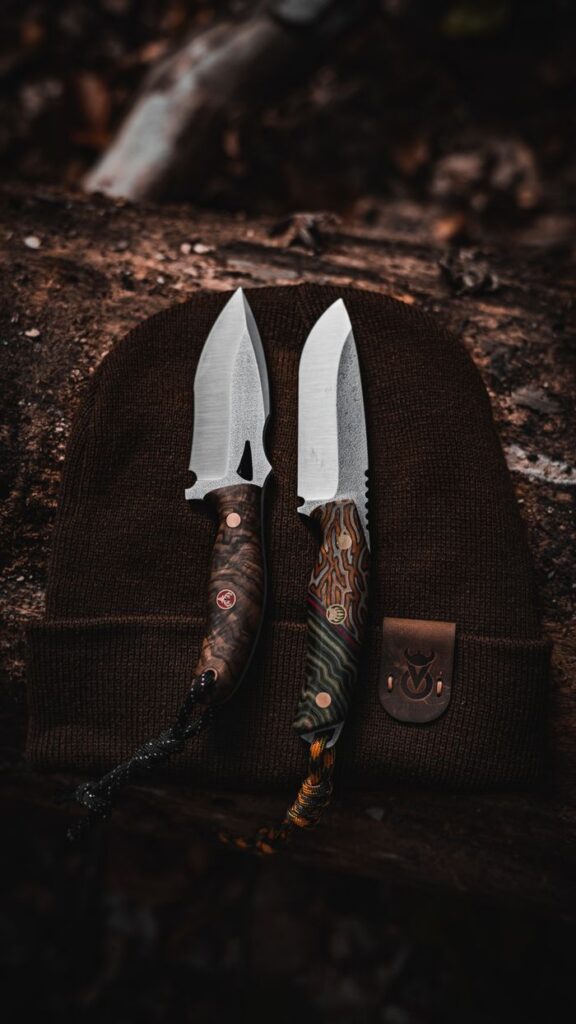 Two handmade knives displayed on a beanie, showcasing intricate handles and craftsmanship.